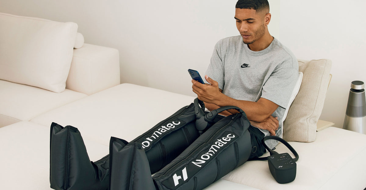 Man sitting on a bed using a smartphone while using the normatec 3 leg system