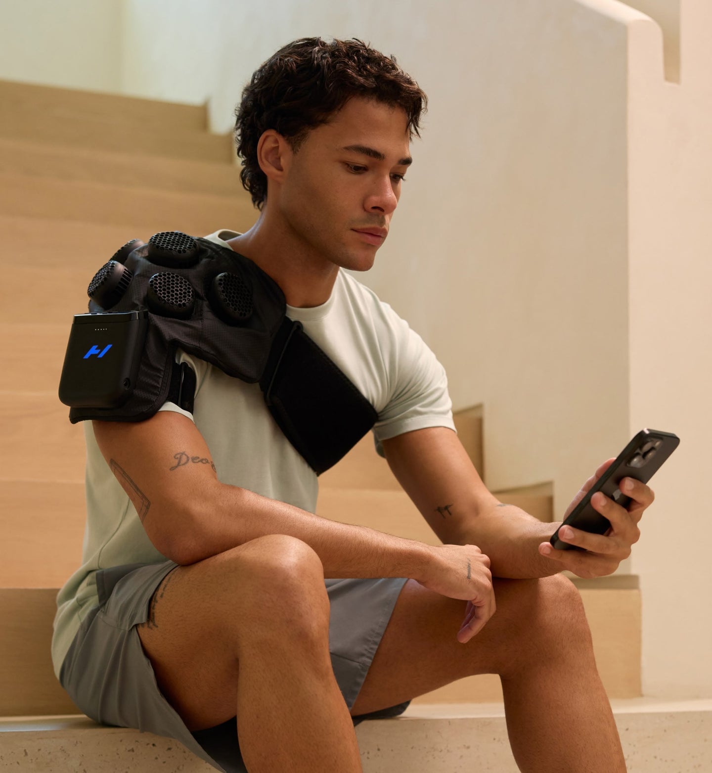 رجل جالس على درجات يستخدم هاتفًا ذكيًا بينما يرتدي Hyperice X 2 على كتفه.