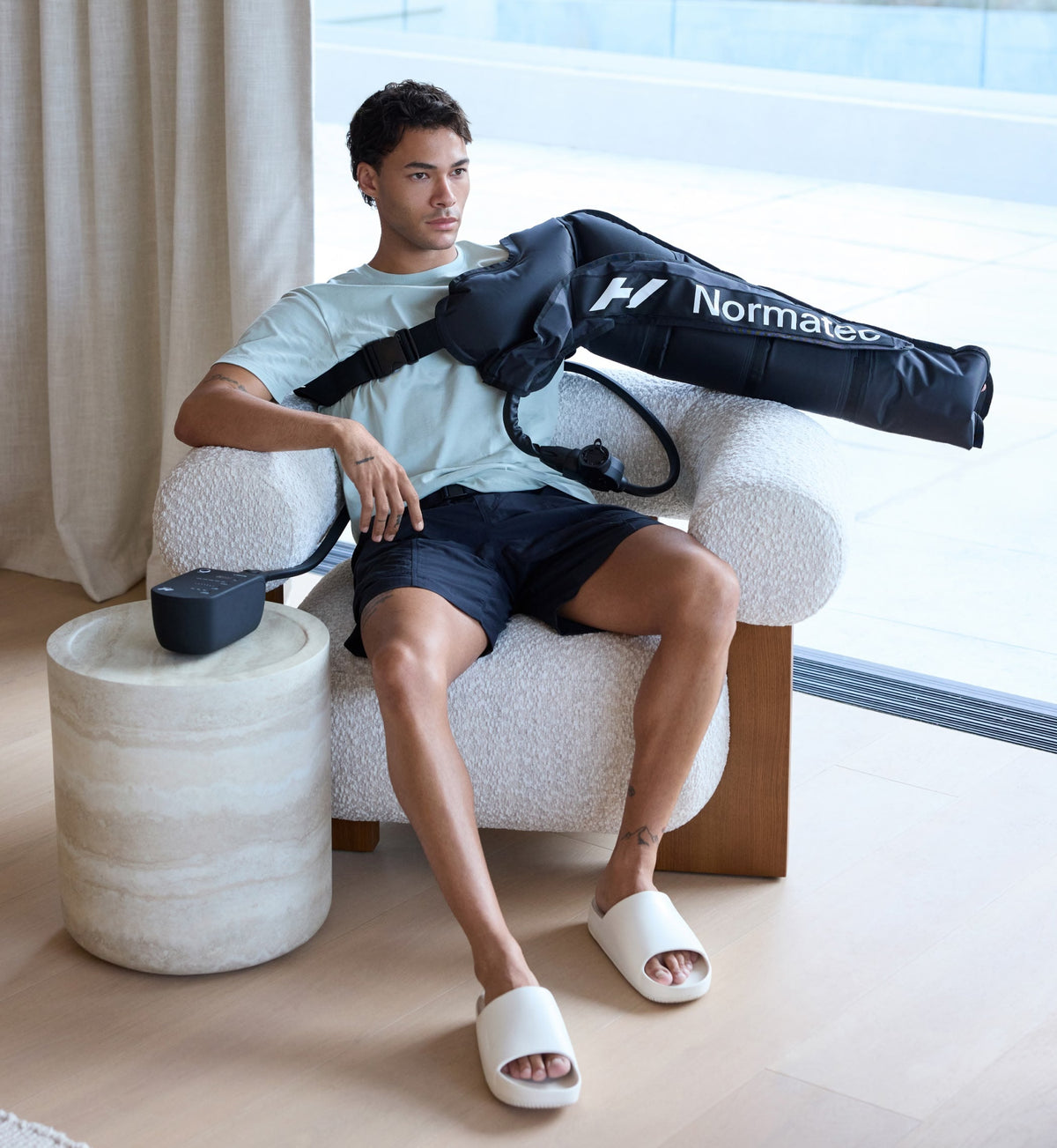 رجل جالس على كرسي مع ملحق ذراع Normatec 3 مثبتًا على ذراعه، في بيئة داخلية عصرية.